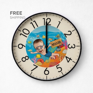 Könnte beinhalten: Runde Wanduhr mit schwarzem Rahmen und cremefarbenem Zifferblatt. Die Uhr zeigt eine Cartoon-Illustration eines Kindes, das unter Wasser schwimmt, umgeben von Fischen und Korallen. Das Kind trägt eine Brille und hält ein Schild mit der Aufschrift "Micah".
