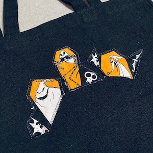May include: Black tote bag with three coffin-shaped patches featuring characters from The Nightmare Before Christmas. The patches have orange, white, and black details. The bag has two black handles.
