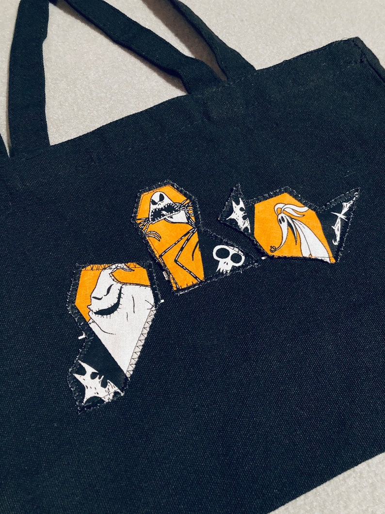 May include: Black tote bag with three coffin-shaped patches featuring characters from The Nightmare Before Christmas. The patches have orange, white, and black details. The bag has two black handles.
