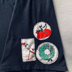 May include: Black t-shirt with three appliqued patches. The patches feature a cat in a bag, a character from The Nightmare Before Christmas, and a wreath with a skull.