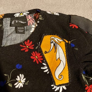 May include: Black and white floral print dress with a yellow coffin-shaped patch featuring a white ghost character. The dress has a black collar and black fabric on the sleeves. The tag on the dress reads "the class™" and "XS (4/5) MADE IN CHINA".