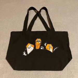 May include: Black tote bag with a Halloween-themed design. The bag features a graphic of characters in orange, white, and black. The tote has two black handles.