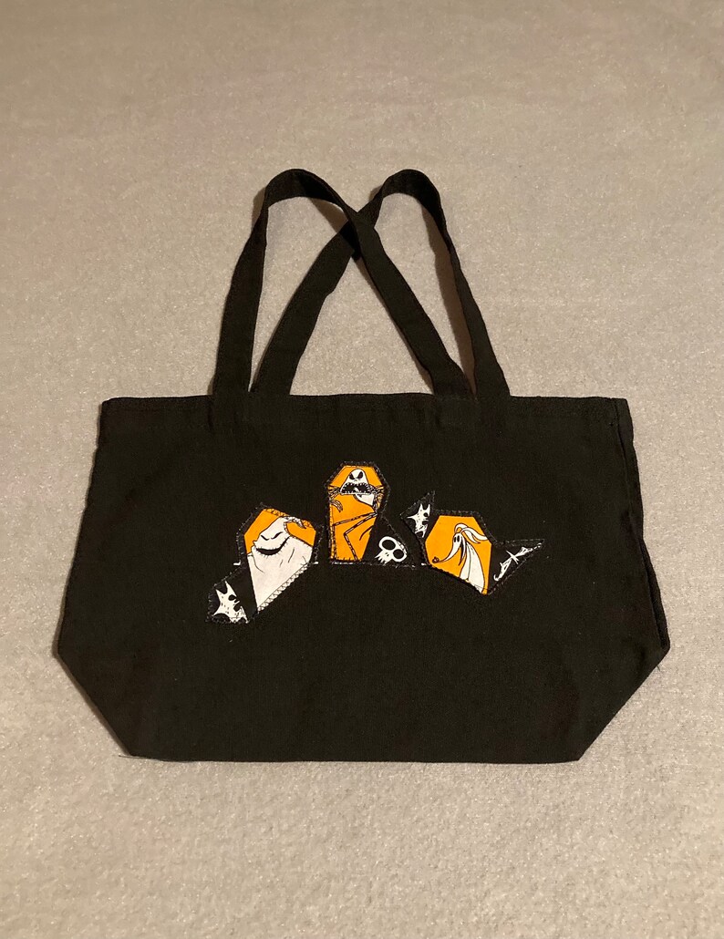 May include: Black tote bag with a Halloween-themed design. The bag features a graphic of characters in orange, white, and black. The tote has two black handles.