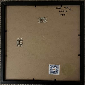 May include: The back of a framed artwork, showcasing a black frame and a tan backing. Metal hanging hardware is visible, along with handwritten text that reads "Jack Toffey 6/9/25 2/250". A QR code sticker is affixed to the lower right corner.