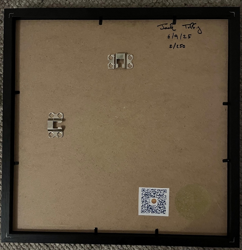May include: The back of a framed artwork, showcasing a black frame and a tan backing. Metal hanging hardware is visible, along with handwritten text that reads "Jack Toffey 6/9/25 2/250". A QR code sticker is affixed to the lower right corner.