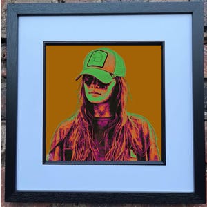 May include: Pop art style portrait of a person wearing a green and orange trucker hat, framed in black. The artwork features a vibrant color palette of orange, green, and purple. The person's long hair and shirt are visible.