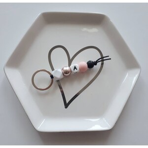 May include: A keychain with a silver ring and a beaded design. The beads are white, rose gold, pink, and black, with the letter "A" on one bead. The keychain rests on a white, hexagonal dish with a heart design.