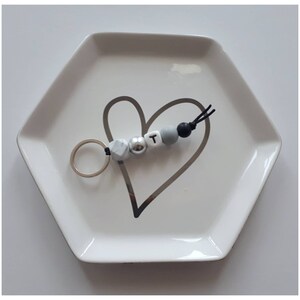 May include: A keychain with a silver ring and a beaded design. The beads include a marbled gray, a silver metallic bead, a white bead with the letter "T", a gray bead, and a black bead. The keychain is on a white hexagonal dish with a gray heart design.