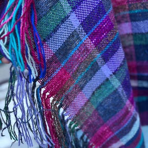 May include: A close-up of a woven textile with a plaid pattern in shades of purple, pink, green, and blue. The textile has a fringe of colorful threads in turquoise, pink, and purple. The fabric appears soft and textured.