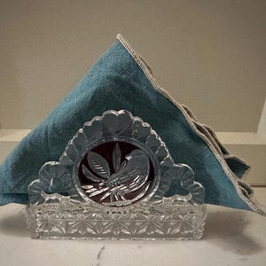 May include: A clear crystal napkin holder with a bird and leaf design, holding a folded teal napkin. The napkin holder has a rectangular base with a diamond pattern and a scalloped top.