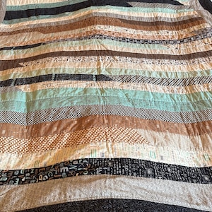 Könnte beinhalten: Eine handgefertigte Steppdecke mit horizontalen Streifen in verschiedenen Farben und Mustern. Die Steppdecke hat Streifen aus hellblauem, braunem, schwarzem und cremefarbenem Stoff. Einige Streifen haben gedruckten Text oder geometrische Muster. Die Steppdecke liegt auf einem hölzernen Bettrahmen.