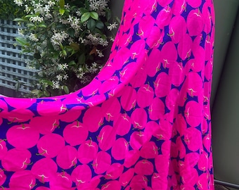 May include: A floor-length wrap skirt featuring a vibrant pattern of bright pink apples set against a deep navy blue background. The skirt has a flowing silhouette with a side closure and a hemline that drapes naturally, showcasing the bold fruit print.