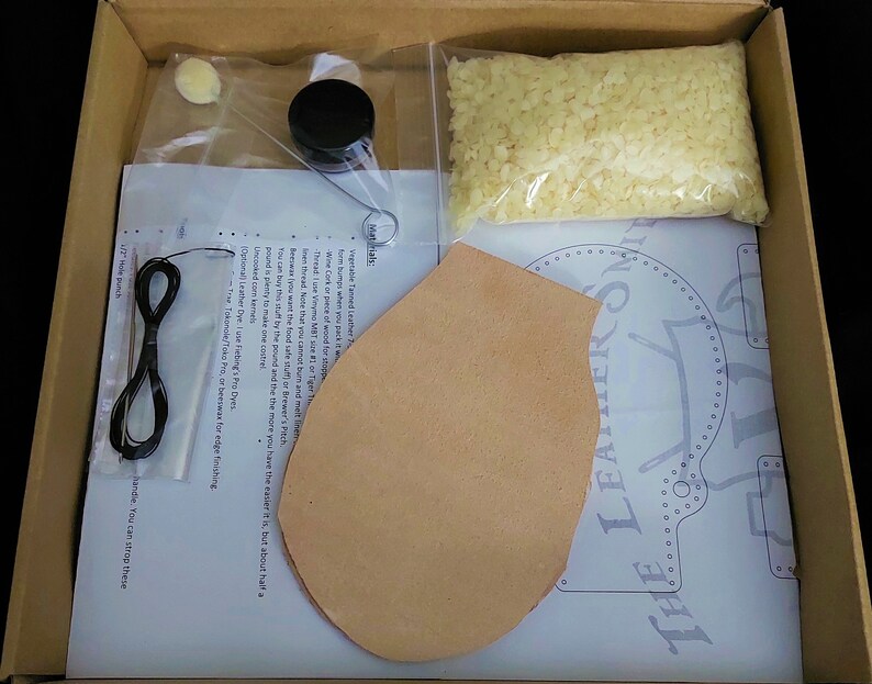 May include: A leatherworking kit with a piece of leather, a pattern, a spool of thread, a small jar of beeswax, a small bag of beads, and instructions. The kit is in a cardboard box.