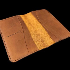 Puede incluir: Una cartera de cuero marrón con un diseño de solapa y un interior de ante. La cartera tiene múltiples ranuras para tarjetas y un compartimento para billetes.