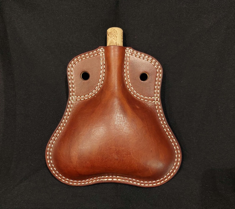 May include: A brown leather flask with a cork stopper and white stitching. The flask has two holes on the top for carrying.