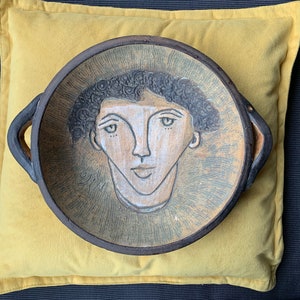 Könnte beinhalten: Ein Keramikteller mit einem handgemalten Porträt einer Person mit dunklen Haaren und einem weißen Gesicht. Der Teller hat zwei Griffe und einen braunen Rand.