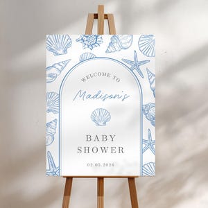 Puede incluir: Un letrero de bienvenida para baby shower sobre un caballete de madera. El letrero es blanco con ilustraciones de conchas marinas azules y el texto "Welcome to Madison's Baby Shower" con la fecha 02.05.2026.