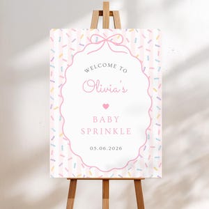 May include: A rectangular welcome sign on a wooden easel featuring a light pink and white vertical striped background with colorful sprinkles. A central white scalloped frame contains the text: WELCOME TO Olivia's BABY SPRINKLE 05.06.2026.
