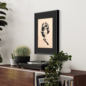 Op de afbeelding: Zwart-wit portret van een vrouw met kort haar, ingelijst in een zwart frame en gemonteerd op een houten paneel. Het portret wordt tentoongesteld op een plank met een platenspeler en planten.
