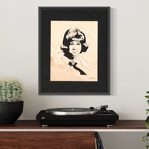 Op de afbeelding: Een zwart-wit portret van een vrouw met kort haar, ingelijst in een zwart frame en hangend aan een witte muur. Het portret is op een houten achtergrond. Een draaitafel staat op een houten plank onder het portret.