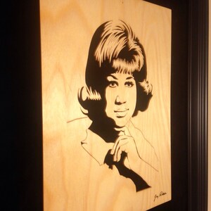 Op de afbeelding: Een zwart-wit houten uitsnijding portret van Aretha Franklin, een beroemde zangeres, met haar kenmerkende kapsel en een hand op haar kin. De uitsnijding is ingelijst in een zwart frame.