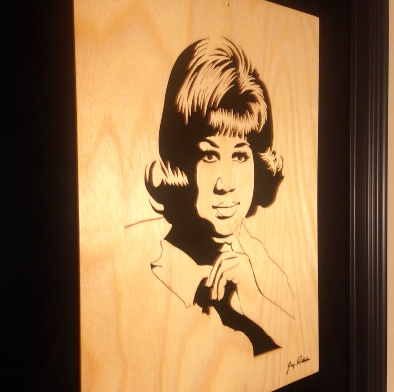 Op de afbeelding: Een zwart-wit houten uitsnijding portret van Aretha Franklin, een beroemde zangeres, met haar kenmerkende kapsel en een hand op haar kin. De uitsnijding is ingelijst in een zwart frame.