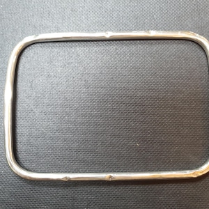 May include: A silver-colored, rectangular metal frame with rounded corners. The frame has a slightly textured surface and appears to be handmade. The object is set against a dark background, highlighting its shape and metallic sheen.