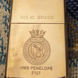 May include: A rectangular, gold-colored lighter made of solid brass. The lighter features the text "SOLID BRASS" at the top and an engraved emblem with the text "PENELOPE" and "HMS PENELOPE F127". The lighter is closed and sits on a blue and white patterned surface.