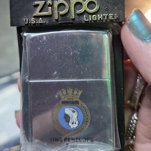 May include: A silver Zippo lighter in its original packaging. The lighter features a crest with a blue and white design and the text "HMS Penelope." The packaging reads "Zippo Lighter U.S.A." and "Lifetime Guarantee."