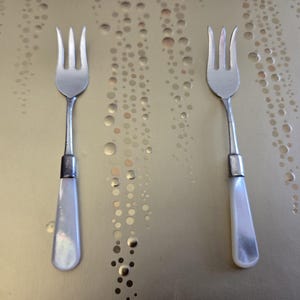 May include: Two antique silver forks with mother-of-pearl handles. The forks have three tines and a decorative silver band where the handle meets the fork. The handles are pearlescent white. The forks are displayed on a gold surface.