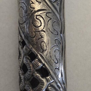 May include: An elaborately decorated silver-coloured metal object, featuring detailed floral and scroll patterns. The cylindrical form has a pointed top and a decorative base, with openwork revealing a dark interior.