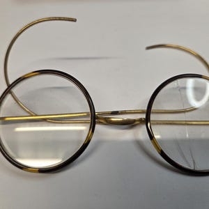 May include: Round eyeglasses with a gold-tone metal frame and clear lenses. The rims are dark brown with a tortoise shell pattern. The earpieces are curved and extend from the frame. The glasses are designed for vision correction.