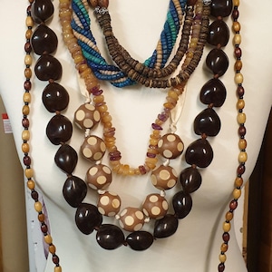 May include: A collection of necklaces displayed on a mannequin. The necklaces feature various designs, including large dark brown beads, dotted beads, and colorful, braided strands. The color palette includes browns, tans, blues, and rainbow hues.