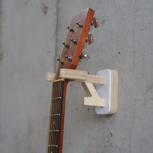 May include: Acoustic guitar mounted on a wall using a wooden guitar holder. The guitar features a brown wooden headstock and neck, with silver tuning pegs. The holder is made of light-coloured wood and white backing, mounted on a grey concrete wall.