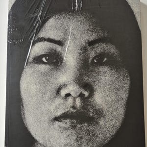 May include: A black and white book cover featuring a close-up portrait of a person's face. The image is grainy, with a high contrast between light and dark areas. The book is sealed in clear plastic.