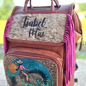 May include: Brown leather backpack with a cowgirl graphic on the front pocket. The backpack features a cowhide panel with the name "Isabel Mae" and pink fringe accents. The graphic depicts a cowgirl riding a horse.