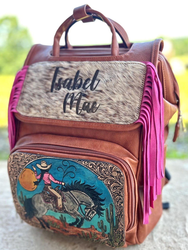 May include: Brown leather backpack with a cowgirl graphic on the front pocket. The backpack features a cowhide panel with the name "Isabel Mae" and pink fringe accents. The graphic depicts a cowgirl riding a horse.