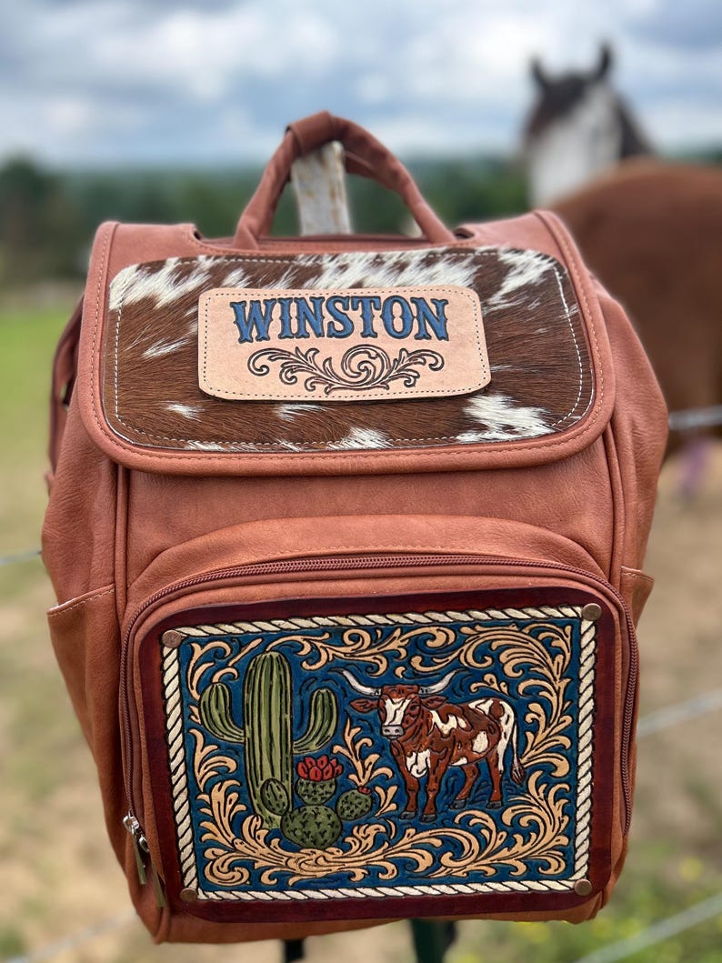 May include: A brown leather backpack with a cowhide flap and the name "WINSTON" in blue lettering. The front features a detailed illustration of a cactus and a longhorn steer. The bag has a top handle and side pockets.
