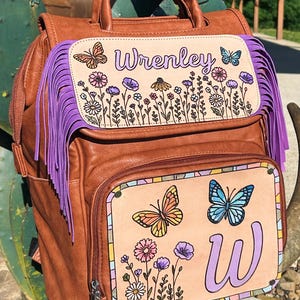 May include: Brown leather backpack with purple fringe and hand-painted floral and butterfly designs. The backpack features the name "Wrenley" and the letter "W". The bag has a top handle and adjustable straps.