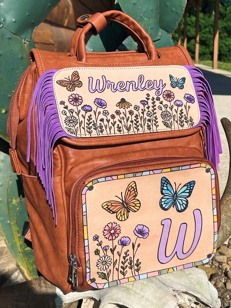 May include: Brown leather backpack with purple fringe and hand-painted floral and butterfly designs. The backpack features the name "Wrenley" and the letter "W". The bag has a top handle and adjustable straps.