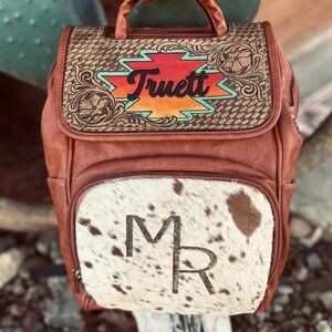 May include: Brown leather backpack with a cowhide front pocket. The flap features the name "Truett" in a colorful design. The front pocket has the initials "MR". The bag has a top handle and side pockets.