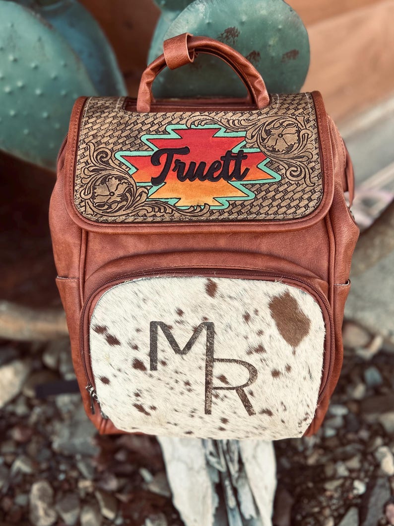 May include: Brown leather backpack with a cowhide front pocket. The flap features the name "Truett" in a colorful design. The front pocket has the initials "MR". The bag has a top handle and side pockets.