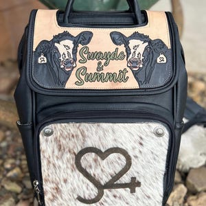 May include: Black and tan backpack with a cowhide front panel. The top flap features two cow illustrations and the text "Swayde & Summit." The front panel has a heart-shaped logo. Includes a top handle and adjustable straps.