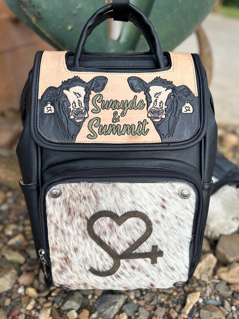 May include: Black and tan backpack with a cowhide front panel. The top flap features two cow illustrations and the text "Swayde & Summit." The front panel has a heart-shaped logo. Includes a top handle and adjustable straps.