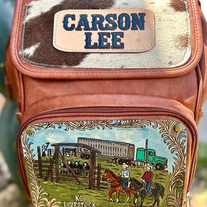 May include: Brown leather backpack with a cowhide panel and the name "CARSON LEE" in blue lettering. The front pocket features a painted scene of cowboys on horseback, a truck, and the text "KG LIVESTOCK".