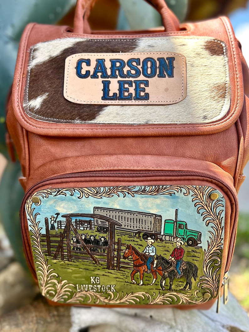 May include: Brown leather backpack with a cowhide panel and the name "CARSON LEE" in blue lettering. The front pocket features a painted scene of cowboys on horseback, a truck, and the text "KG LIVESTOCK".