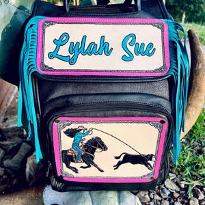 May include: Black leather backpack with turquoise fringe and a pink and cream-colored rectangular patch that reads "Lylah Sue" in turquoise script. The lower patch depicts a cowgirl roping a calf.