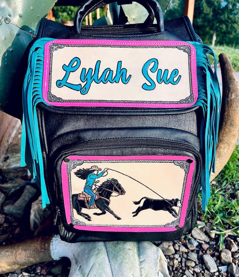 May include: Black leather backpack with turquoise fringe and a pink and cream-colored rectangular patch that reads "Lylah Sue" in turquoise script. The lower patch depicts a cowgirl roping a calf.