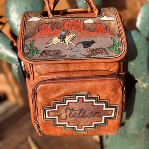 May include: A brown leather backpack with a western-themed design. The top flap features a painted desert scene with a cowboy on a horse roping a steer. The front pocket has the name "Stetson" in a decorative frame.