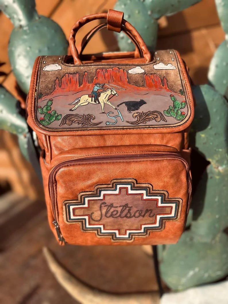 May include: A brown leather backpack with a western-themed design. The top flap features a painted desert scene with a cowboy on a horse roping a steer. The front pocket has the name "Stetson" in a decorative frame.